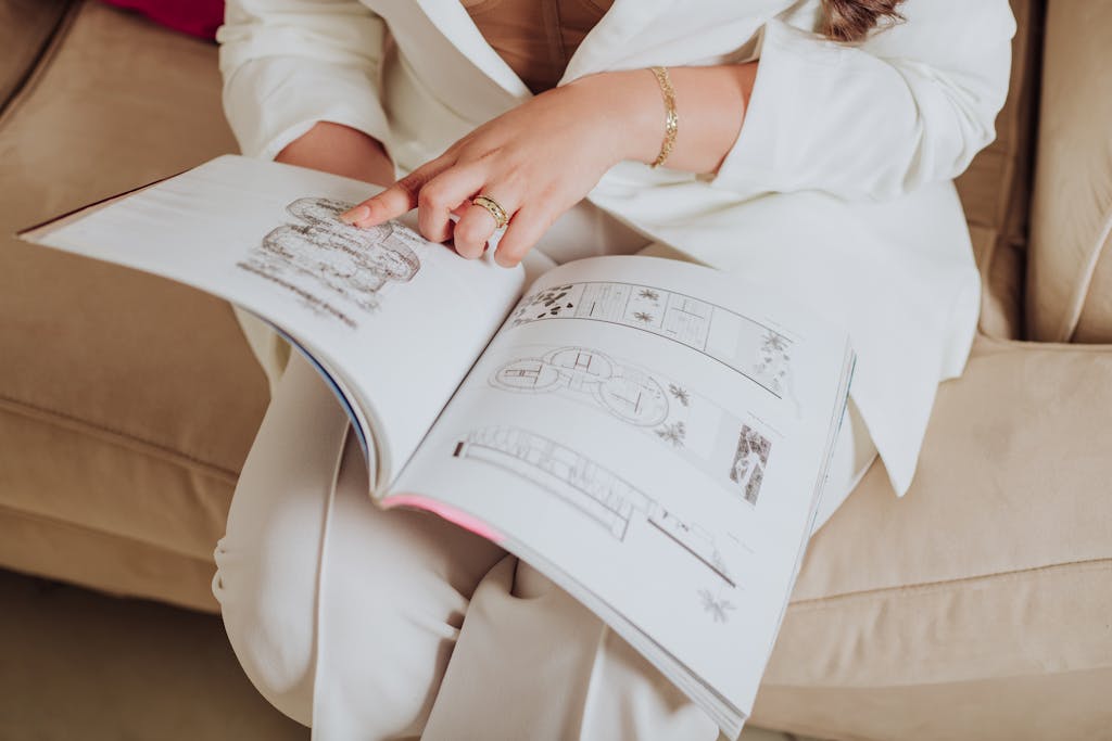 A woman in elegant attire reviewing architectural designs in a magazine while sitting on a sofa.